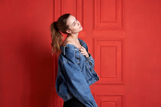 Potrtait de sensual garota sexy com penteado fofo e olhos fechados, vestindo uma jaqueta jeans da moda e calça preta na parede vermelha. Estúdio filmado em fundo vermelho.