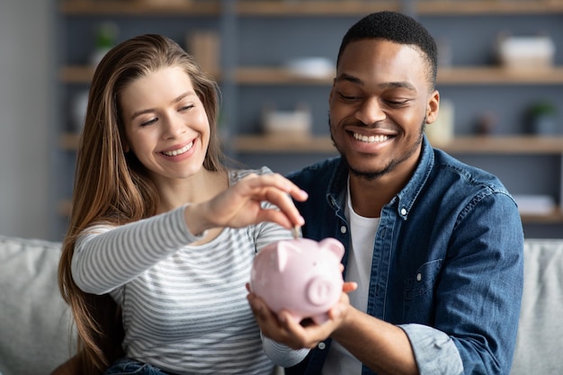 Foto poupança familiar. jovem casal interracial colocando moedas no cofrinho em casa enquanto sentados juntos no sofá na sala de estar, amantes multiculturais alegres planejando orçamento comum juntos, closeup