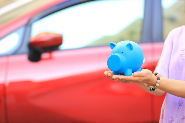 Poupar dinheiro e empréstimos para o conceito de carro, jovem mulher segurando o porquinho azul com pé no estacionamento, auto negócio