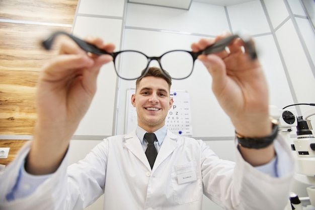 POV-Aufnahme eines lächelnden Augenoptikers, der eine Brille auf einen nicht erkennbaren Patienten setzt