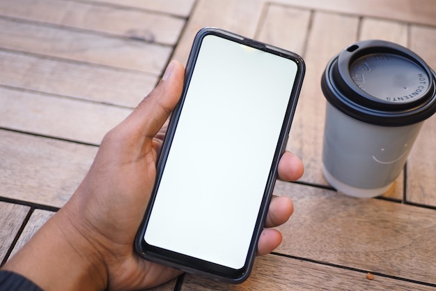 Pov-Aufnahme, wie man ein Smartphone mit weißem Bildschirm und Papierkaffee auf dem Tisch hält