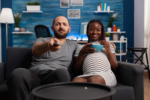 Pov de grávidas interraciais, olhando para a câmera assistindo televisão na sala de estar. parceiros de raça mista com gravidez relaxando enquanto comem pipoca e usam o controle remoto da tv