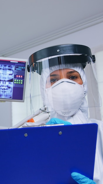 Pov de paciente recebendo tratamento dentário no consultório do dentista com novo normal, médico tomando notas na área de transferência antes de examinar. Stomatolog usando equipamento de segurança contra coronavírus durante a verificação de cuidados de saúde