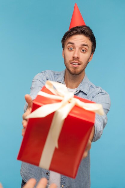 POV mano dando caja de regalo roja envuelta a hombre barbudo con cono de fiesta en la cabeza, tipo con camisa de mezclilla mirando sorpresa de regalo de cumpleaños con deleite. Disparo de estudio interior aislado sobre fondo azul.