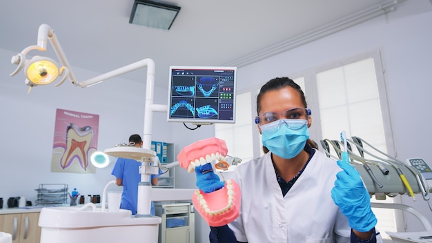 Foto pov paciente de dentista ensinando mulher correta limpeza de dentes em consultório odontológico usando acessório de esqueleto. stomatolog usando máscara de proteção pedindo apresentação de objeto médico de dentes