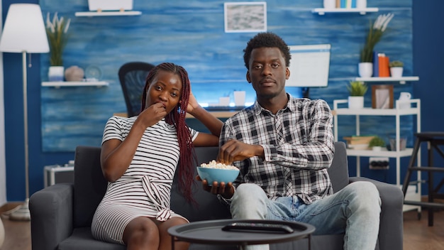 POV de una pareja afroamericana viendo la televisión mientras come palomitas de maíz del tazón. Amantes negros sentados en el sofá en la sala de estar mientras miran a la cámara. Jóvenes relajándose en casa