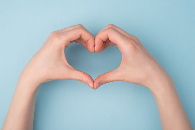 Pov superior por encima de la cabeza vista de cerca la foto de las manos haciendo forma de corazón aislado sobre fondo de color azul pastel