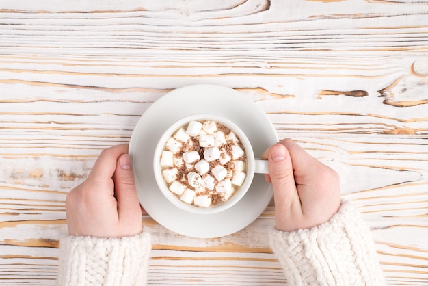 Pov-Top über Overhead Nahaufnahme Foto von weiblichen Händen, die eine Tasse leckeres Getränk mit weichem Zuckermarshmallow auf Holzhintergrund halten