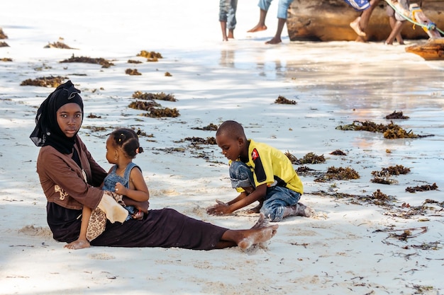 Povo da África. Mulher africana com crianças senta-se na areia da praia. Zanzibar.