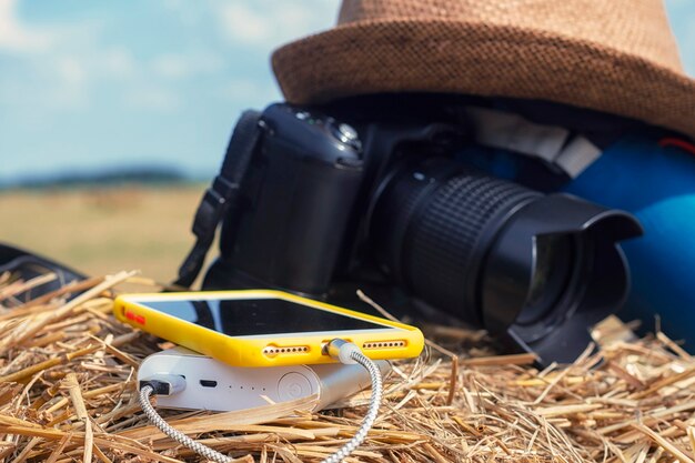 Power Bank carga un teléfono inteligente en el fondo de una cámara con una bolsa y un sombrero sobre un fondo de heno en la naturaleza. Cargador de viaje portátil.