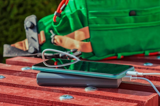 Powerbank lädt ein Smartphone auf einer Bank mit einer grünen Tasche auf. Tragbares Ladegerät für Gadgets