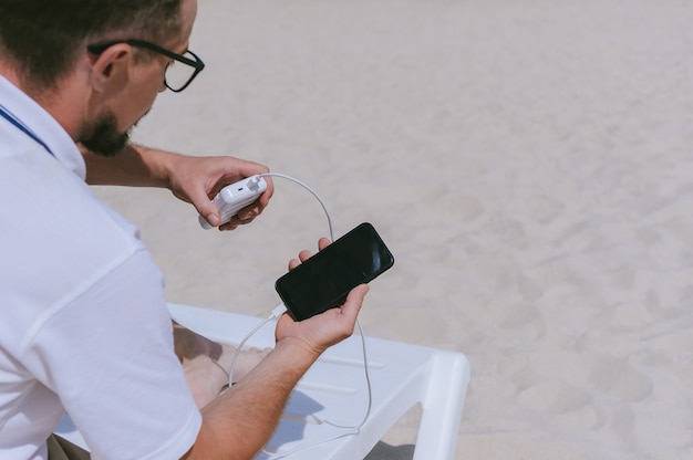 Powerbank lädt ein Smartphone in den Händen eines Mannes am Strand auf. Vor dem Hintergrund von Sand.