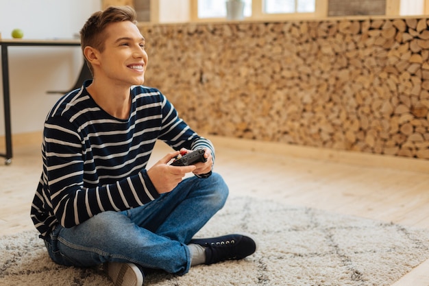 Foto practicando juegos. hermoso inspirado joven rubio sonriendo y sosteniendo un control remoto para juegos mientras está sentado en el piso