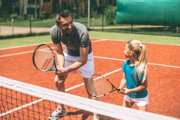 Practicando tenis. Padre alegre en ropa deportiva enseñando a su hija a jugar al tenis mientras ambos están de pie en la cancha de tenis