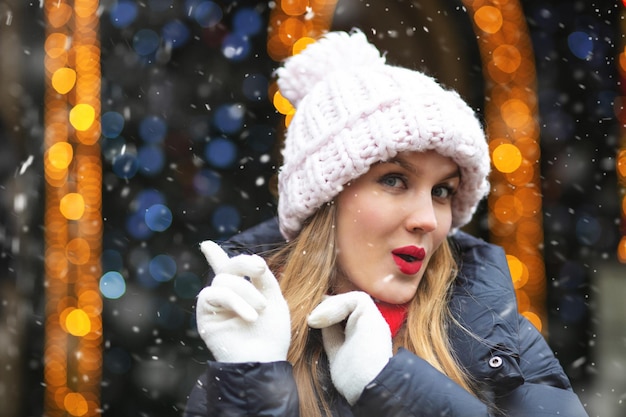 Prächtige blonde Frau trägt Strickmütze, die bei Schneefall auf der Wintermesse geht. Leerer Platz für Text