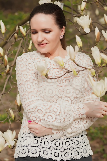 Prächtige brünette Frau mit nacktem Make-up, die Spitzenbluse trägt und in der Nähe der blühenden Magnolienblumen posiert