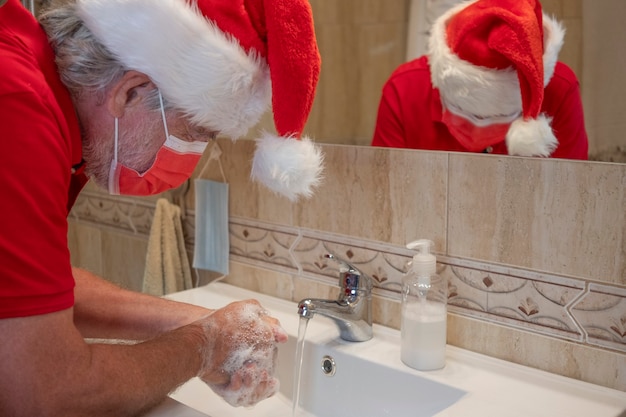 Prävention der Coronavirus-Pandemie. Mann wäscht sich zu Hause die Hände mit einer chirurgischen Maske wegen Coronavirus, mit einer Weihnachtsmütze auf dem Kopf. Fokus auf Schaum