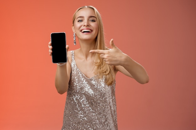 Prahlerische stolze charmante elegante blonde Frau im stilvollen Abendkleid zeigen Smartphone-Anzeige stolz zeigend Handy-Bildschirm lächelnd zeigt Foto Freund, stehend roten Hintergrund.