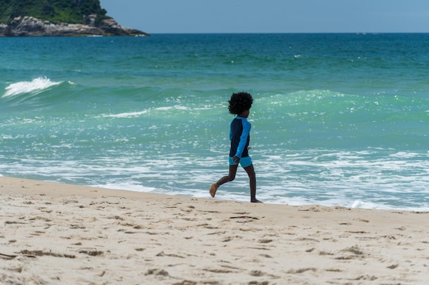 Praia de Grumari perto da Barra da Tijuca no Rio de Janeiro, Brasil. Dia ensolarado com céu azul, água clara. Colinas e natureza ao redor. Criança negra de costas, brincando na areia.