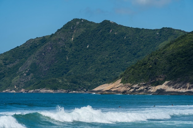 Praia de Grumari perto da Barra da Tijuca no Rio de Janeiro, Brasil. Dia ensolarado com céu azul, águas claras e pequenas ondas. Colinas e natureza ao redor.