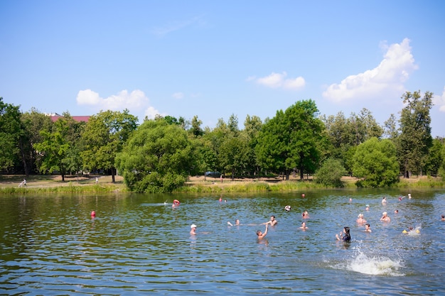 Praia fluvial ou lacustre com pessoas nadando entre árvores verdes, dia de sol.