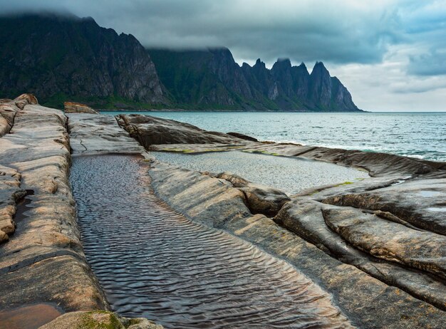 Praia pedregosa com banhos de maré em Ersfjord, Senja, Noruega. Costa de noite de dia polar de verão. Os dentes do dragão balançam longe.