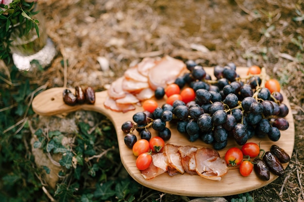 Prancha de madeira para cortar com um cacho de uvas, presunto, tomate e tâmaras na natureza