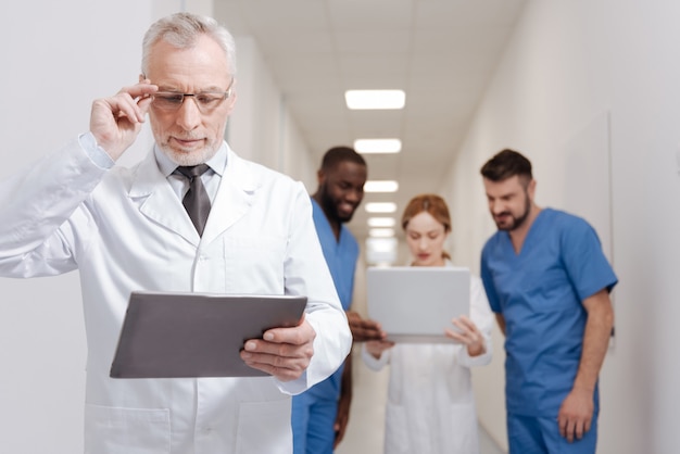 Foto praticando uma nova abordagem. sênior barbudo pediatra envolvido aproveitando o trabalho na clínica e trabalhando com um tablet, enquanto outros colegas usam o laptop em segundo plano