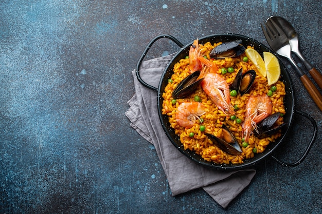 Prato clássico da Espanha, paella de frutos do mar na tradicional panela na vista superior do fundo de pedra azul. Paella espanhola com camarões, pinças, mexilhões, ervilhas e rodelas de limão fresco de cima, espaço para texto