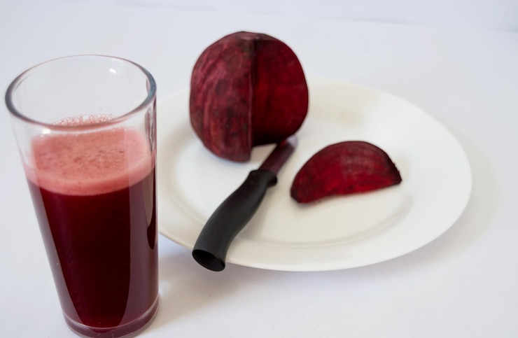 Foto prato com beterraba faltando uma fatia que é a frente no mesmo prato e faca com caldo de beterraba avermelhada e um copo na frente a cena de fundo fica desfocada