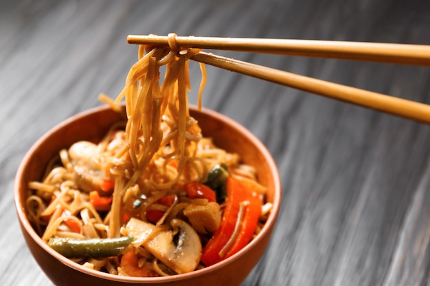 Prato com macarrão de trigo sarraceno com legumes cogumelos carne de frango em um fundo escuro Paus chineses levam soba japonês de uma tigela de barro em um fundo de madeira