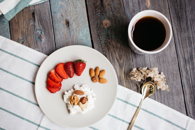 Prato com queijo cottage, morangos e nozes, uma xícara de café e toalhas na mesa de madeira, alimentação saudável, café da manhã