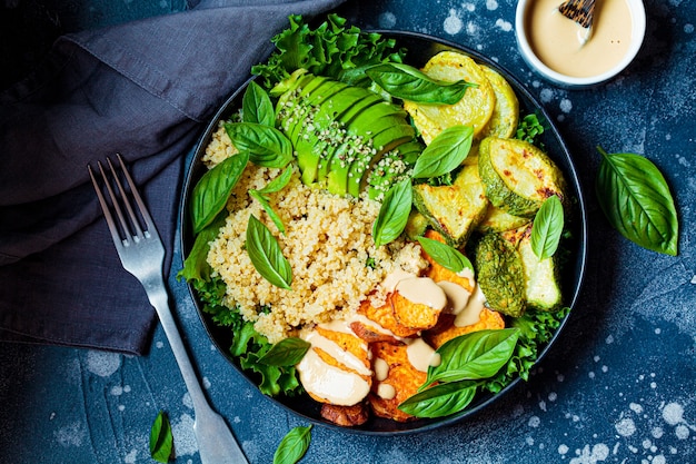 Prato de almoço vegano. salada de quinua com abobrinha assada, batata doce, abacate e molho de tahine em prato preto