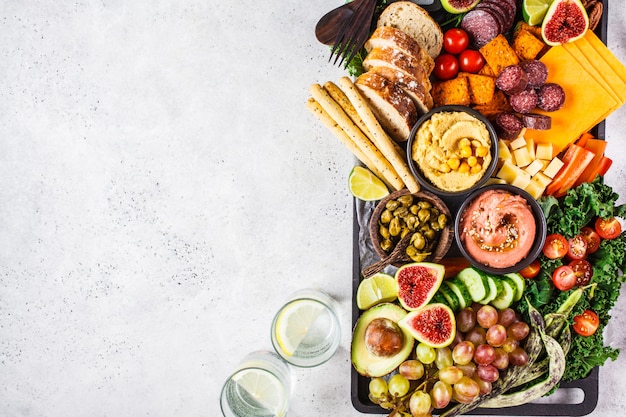 Prato de aperitivos de carne e queijo. Salsicha, queijo, homus, legumes, frutas e pão na bandeja preta.