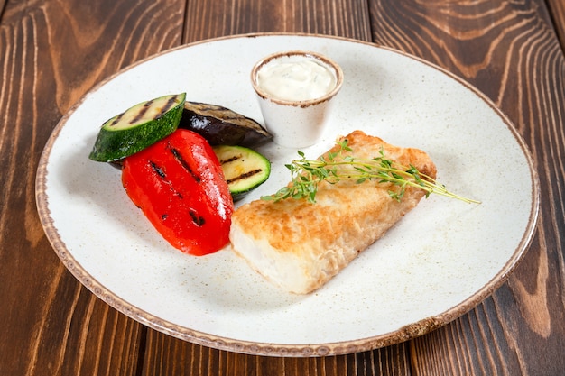 Prato de bacalhau grelhado com legumes e molho branco na mesa de madeira