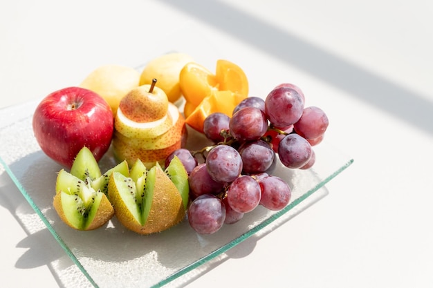 Prato de bagas de frutas cruas laranjas kiwi toranja uvas pêra maçãs no prato na mesa branca