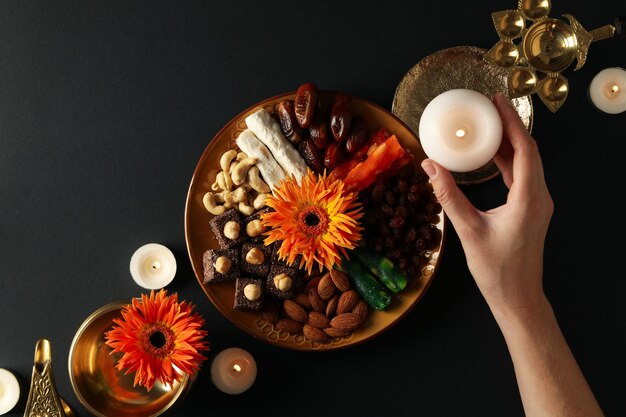 Foto prato de cobre com doces e nozes velas flores e mão em fundo preto vista superior