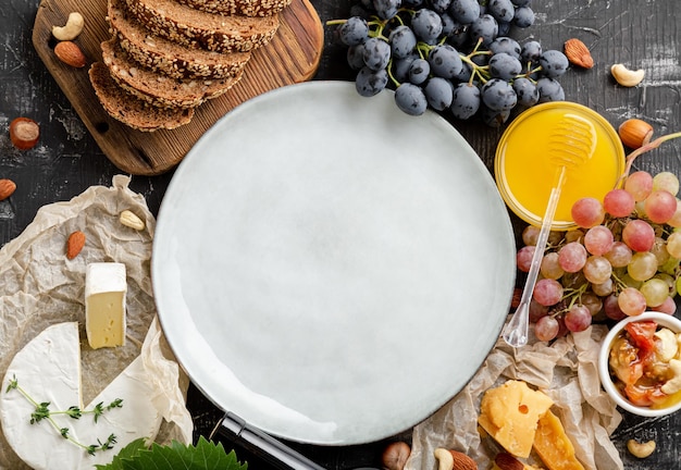 Prato de maquete vazio na mesa de abundância com mantimentos. Prato de maquete servido em moldura feita de comida para o jantar gourmet Produtos pão queijos mel uvas vinho. Vista superior em fundo escuro de concreto.