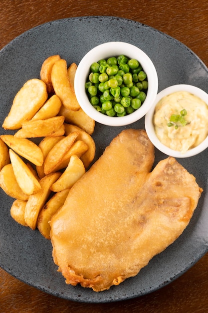 Prato de peixe e batatas fritas com peixe frito fatiado e closeup de mesa de pub com molho tártaro e ervilhas verdes no retrato de vista superior