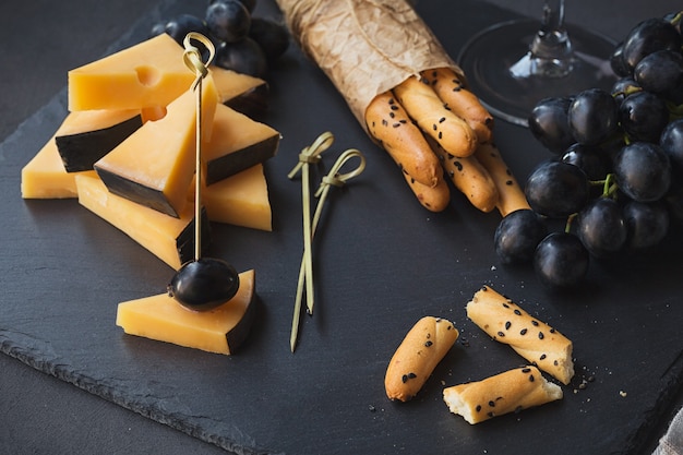 Prato de queijo servido com biscoitos, uvas e copo de vinho branco em fundo escuro. Queijo gouda velho no prato de degustação