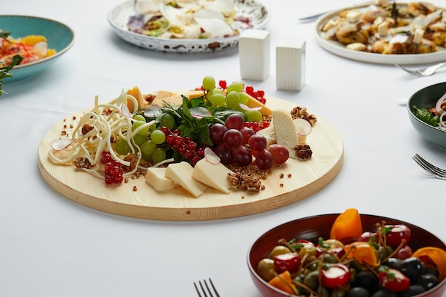 Prato grande de queijos sortidos a meio da mesa com vários petiscos, mix de queijos num prato de madeira como aperitivo principal da mesa.