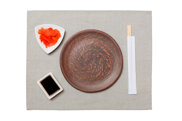 Prato marrom redondo vazio com pauzinhos para sushi, gengibre e molho de soja em fundo de guardanapo cinza. vista superior com espaço de cópia para você projetar.