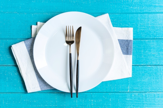 Prato redondo cerâmico branco vazio com toalha de mesa, faca e garfo na mesa de madeira azul.