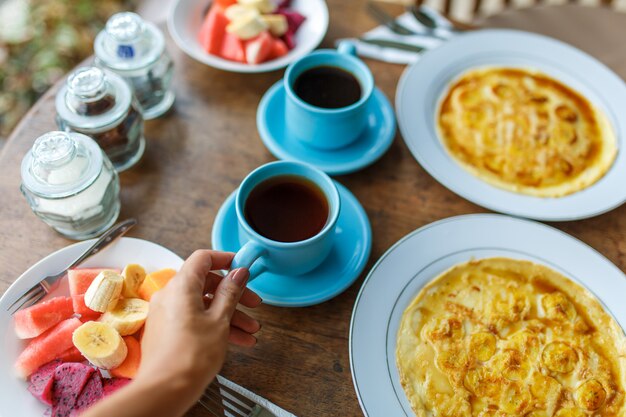 Foto pratos com panquecas de banana, frutas tropicais e duas xícaras de café na mesa de madeira.