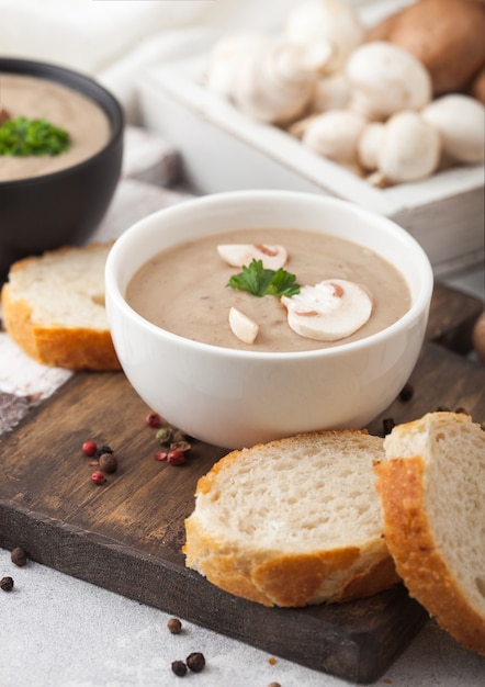 Pratos tigela de cerâmica de sopa cremosa de cogumelos champignon castanha com colher, pimenta e pano de cozinha no fundo branco da cozinha e uma caixa de cogumelos crus.