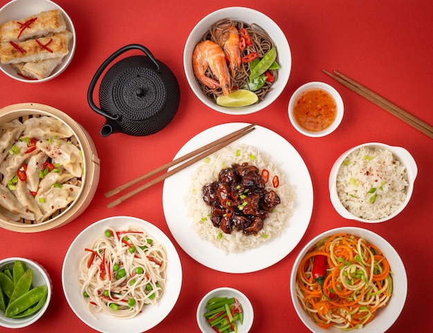 Foto pratos variados da culinária tradicional chinesa em fundo vermelho: frango agridoce com arroz, bolinhos no vapor de bambu, pãezinhos fritos, macarrão e chaleira. conceito de comida asiática, vista de cima