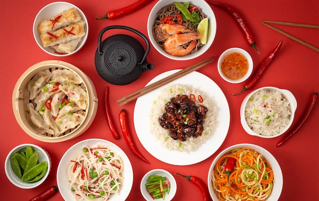 Pratos variados da culinária tradicional chinesa em fundo vermelho: frango agridoce com arroz, bolinhos no vapor de bambu, pãezinhos fritos, macarrão e chaleira. Conceito de comida asiática, vista de cima