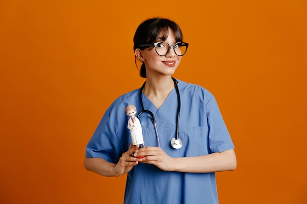 Prazer segurando o brinquedo jovem médica usando estetoscópio uniforme de quinto isolado em fundo laranja