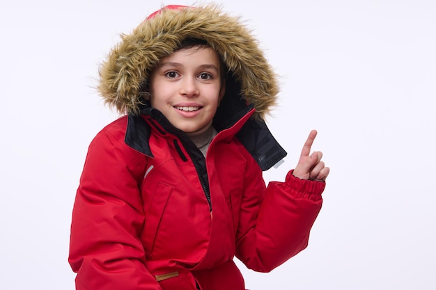 Foto preadolescente alegre guapo, niño caucásico en edad escolar vistiendo una parka roja brillante con capucha, sonriendo sonrisa con dientes mirando a la cámara y apuntando con el dedo en un espacio de copia de fondo blanco