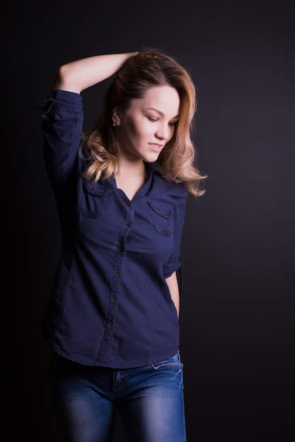 Preciosa modelo joven con pelo largo y rizado viste camisa y jeans, posando sobre un fondo negro